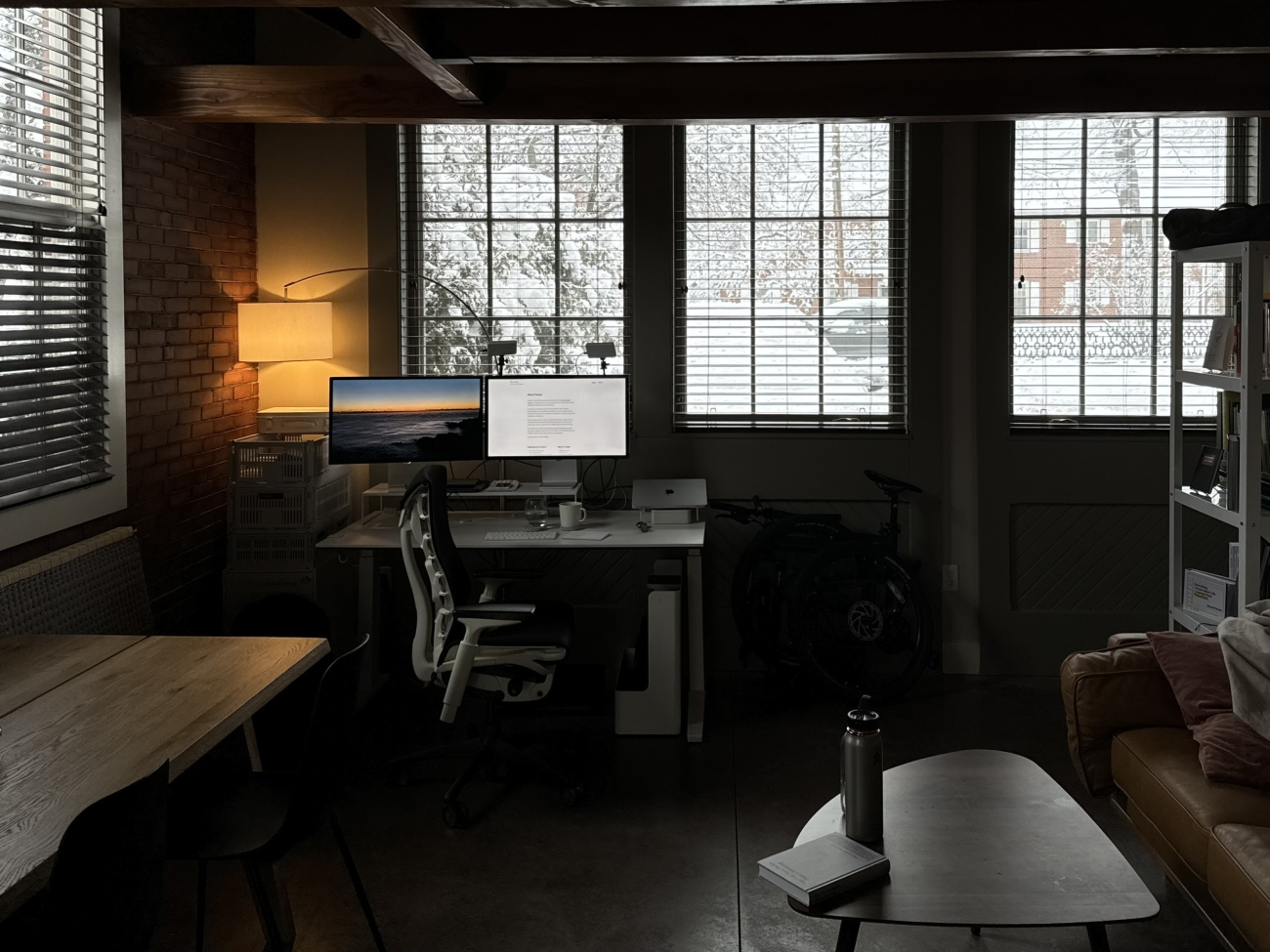 A room in a carriage house apartment with large windows. In the room there is a wooden table, a white desk, an office chair, a bookshelf, and a couch. Outside there is snow nestled on a stand of trees visible through the window.