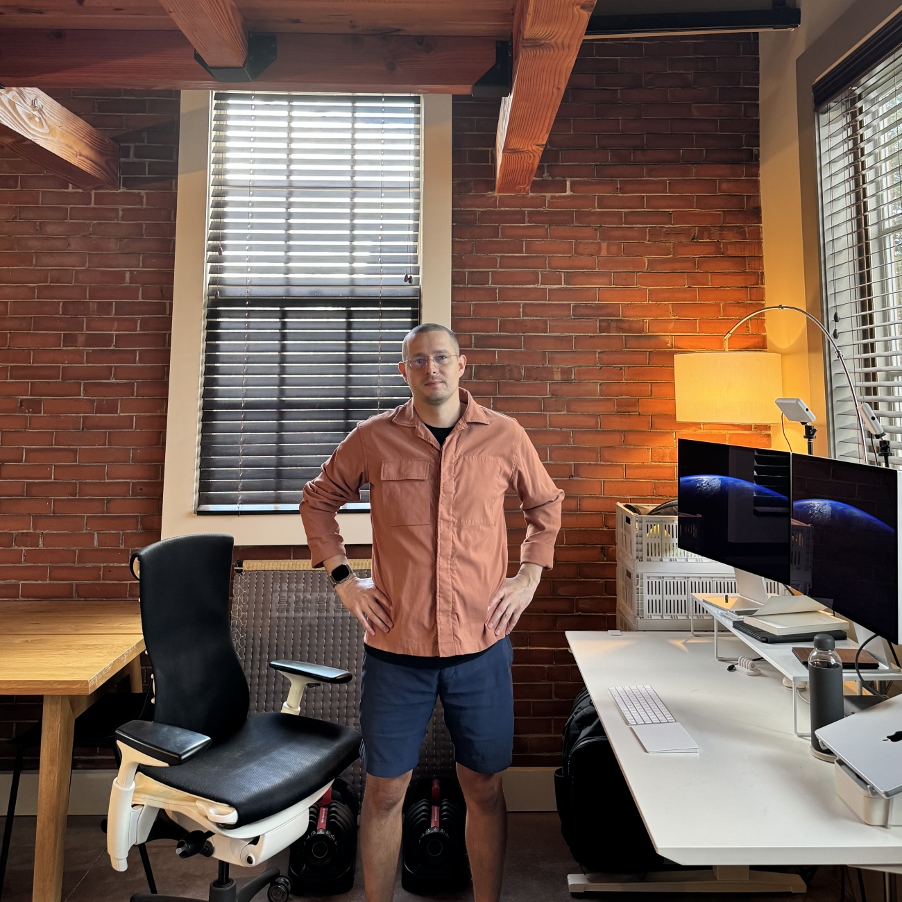 Christian is standing, arms akimbo, between his office chair and desk, looking at the camera. He wears a salmon-colored jacket.